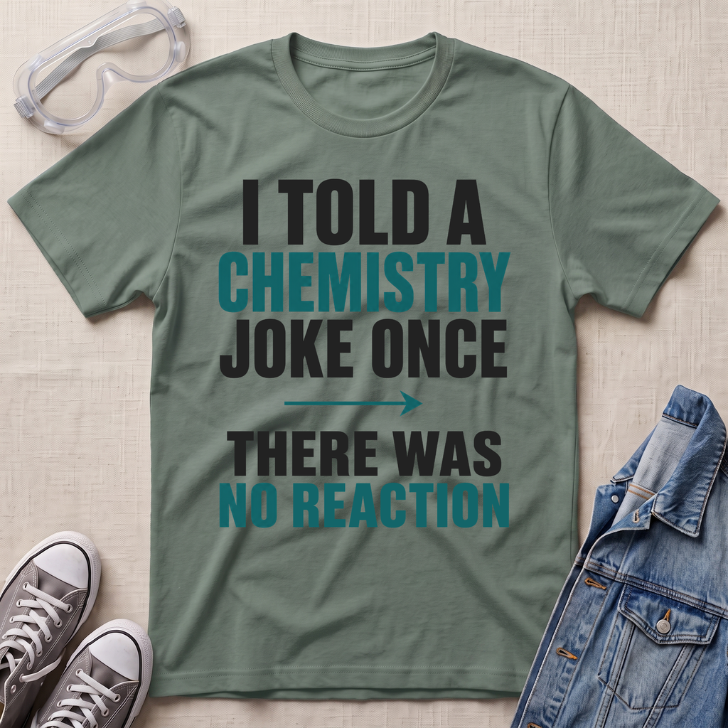 An olive green graphic t-shirt laid flat on a textured beige background, surrounded by styling props including safety goggles, gray sneakers, and a denim jacket. The t-shirt features a printed slogan in black and teal text that reads: 'I TOLD A CHEMISTRY JOKE ONCE THERE WAS NO REACTION'.