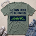 An olive green graphic t-shirt laid flat on a textured beige background, styled with safety goggles, gray sneakers, and a denim jacket. The t-shirt features a dark blue graphic that reads 'QUANTUM MECHANICS' and a retro-style message box in green and blue.