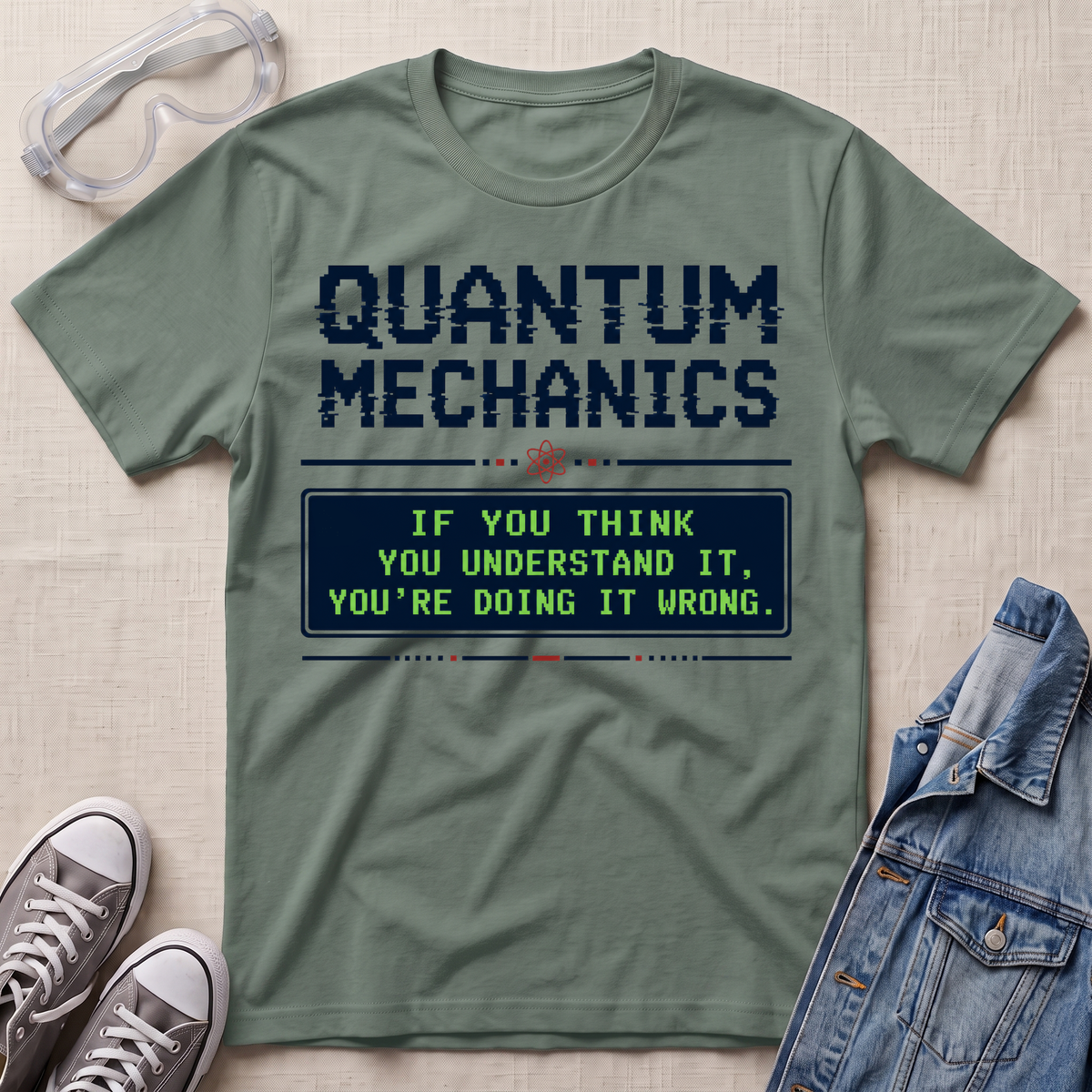 An olive green graphic t-shirt laid flat on a textured beige background, styled with safety goggles, gray sneakers, and a denim jacket. The t-shirt features a dark blue graphic that reads 'QUANTUM MECHANICS' and a retro-style message box in green and blue.