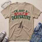 A beige graphic t-shirt laid flat on a textured light background, surrounded by styling props including safety goggles, gray sneakers, and a denim jacket. The t-shirt features the text 'I'M JUST A LITTLE DERIVATIVE' in dark green and red lettering, with the black derivative math formula printed below.