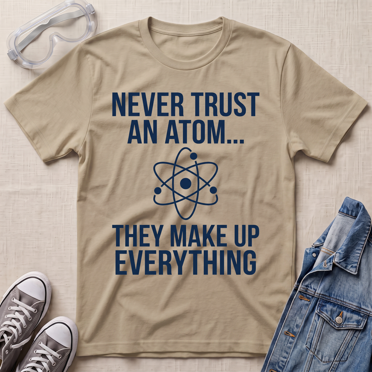 A beige graphic t-shirt with white text reading 'NEVER TRUST AN ATOM.' and 'THEY MAKE UP EVERYTHING' with an image of an atom in between the texts.