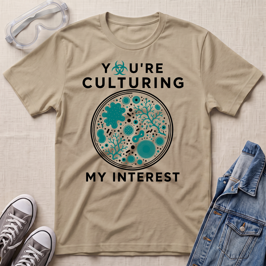 A beige graphic t-shirt laid flat on a textured fabric background, styled with safety goggles, sneakers, and a denim jacket. The t-shirt features a circular microbiology petri dish graphic filled with teal microorganisms and the text 'YOU'RE CULTURING MY INTEREST' in black.