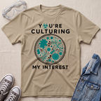 A beige graphic t-shirt laid flat on a textured fabric background, styled with safety goggles, sneakers, and a denim jacket. The t-shirt features a circular microbiology petri dish graphic filled with teal microorganisms and the text 'YOU'RE CULTURING MY INTEREST' in black.