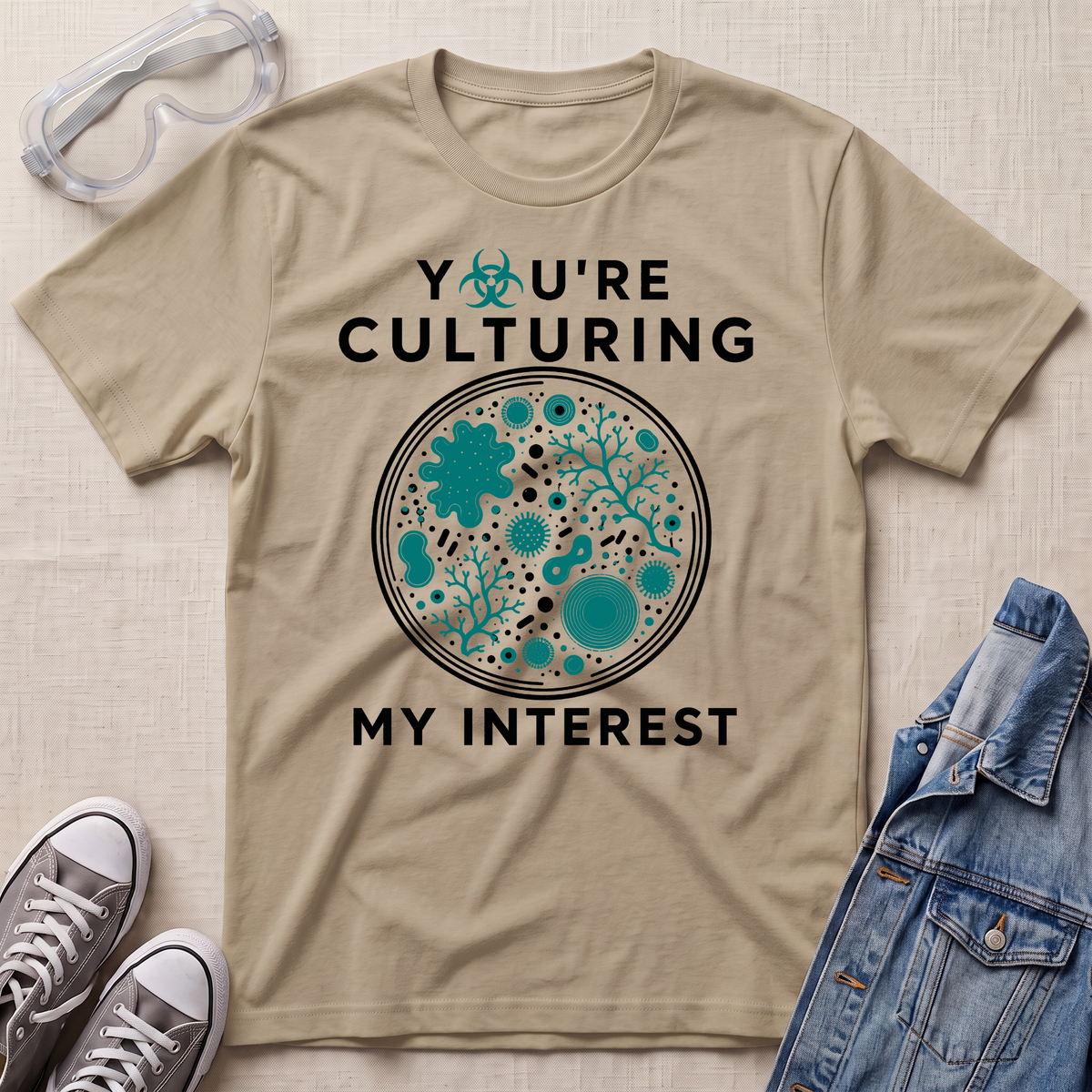 A beige graphic t-shirt laid flat on a textured fabric background, styled with safety goggles, sneakers, and a denim jacket. The t-shirt features a circular microbiology petri dish graphic filled with teal microorganisms and the text 'YOU'RE CULTURING MY INTEREST' in black.
