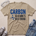 A beige graphic t-shirt laid flat on a textured beige background, styled with safety goggles, gray sneakers, and a denim jacket. The t-shirt features the text 'CARBON IS A GIRL'S BEST FRIEND' in blue and black, with a small blue molecule graphic and a small diamond icon.