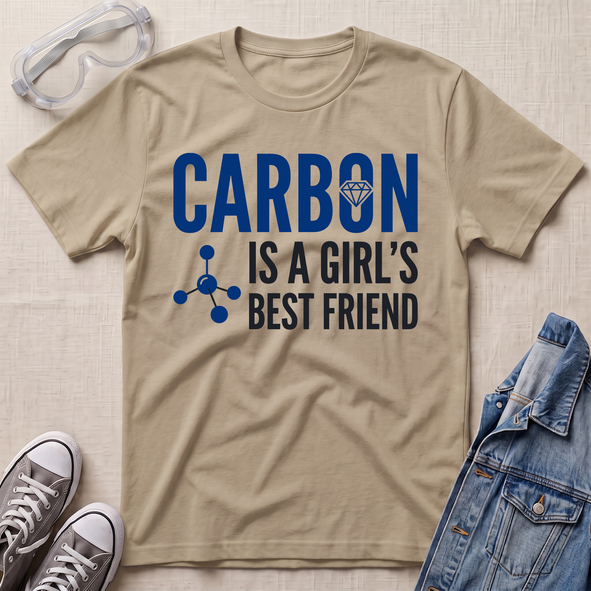 A beige graphic t-shirt laid flat on a textured beige background, styled with safety goggles, gray sneakers, and a denim jacket. The t-shirt features the text 'CARBON IS A GIRL'S BEST FRIEND' in blue and black, with a small blue molecule graphic and a small diamond icon.