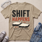 A beige graphic t-shirt laid flat on a textured beige fabric background, styled with safety goggles, gray sneakers, and a blue denim jacket. The t-shirt features a graphic that reads 'SHIFT HAPPENS' in black and red, with illustrations of tectonic plates and the text 'PLATE TECTONICS' below.