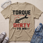 A beige graphic t-shirt laid flat on a textured fabric background, styled with safety glasses, gray sneakers, and a blue denim jacket. The t-shirt features a black and red graphic that reads 'TORQUE DIRTY TO ME' with a wrench and gear illustration.