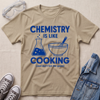 A beige graphic t-shirt laid flat on a textured beige fabric background, styled with safety goggles, gray sneakers, and a blue denim jacket. The t-shirt features a blue graphic that reads 'CHEMISTRY IS LIKE COOKING (Just don't lick the spoon)' with illustrations of a beaker and a mixing bowl.