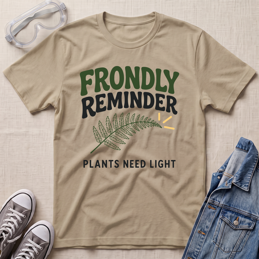 A beige sand graphic t-shirt laid flat on a textured fabric background, styled with safety goggles, gray sneakers, and a denim jacket. The t-shirt features the text 'FRONDLY REMINDER' in green and black, a green fern leaf, and 'PLANTS NEED LIGHT' in black.