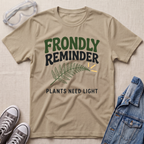 A beige sand graphic t-shirt laid flat on a textured fabric background, styled with safety goggles, gray sneakers, and a denim jacket. The t-shirt features the text 'FRONDLY REMINDER' in green and black, a green fern leaf, and 'PLANTS NEED LIGHT' in black.
