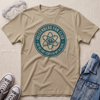 A beige graphic t-shirt featuring a circular 'Heisenberg Fan Club' design in teal, styled in a flat-lay with safety goggles, sneakers, and a denim jacket on a textured beige background.