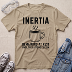 A beige graphic t-shirt laid flat on a textured fabric background, surrounded by styling props including safety goggles, gray sneakers, and a blue denim jacket. The t-shirt features a black graphic of a steaming coffee mug with the text 'INERTIA REMAINING AT REST UNTIL THE CAFFEINE KICKS IN'.