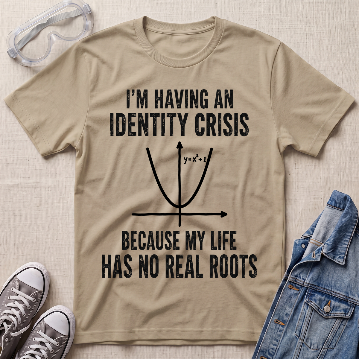 A beige graphic t-shirt laid flat on a textured fabric background, surrounded by styling props including safety goggles, gray sneakers, and a blue denim jacket. The t-shirt features a black printed graphic of a parabola with the text 'I'M HAVING AN IDENTITY CRISIS BECAUSE MY LIFE HAS NO REAL ROOTS'.