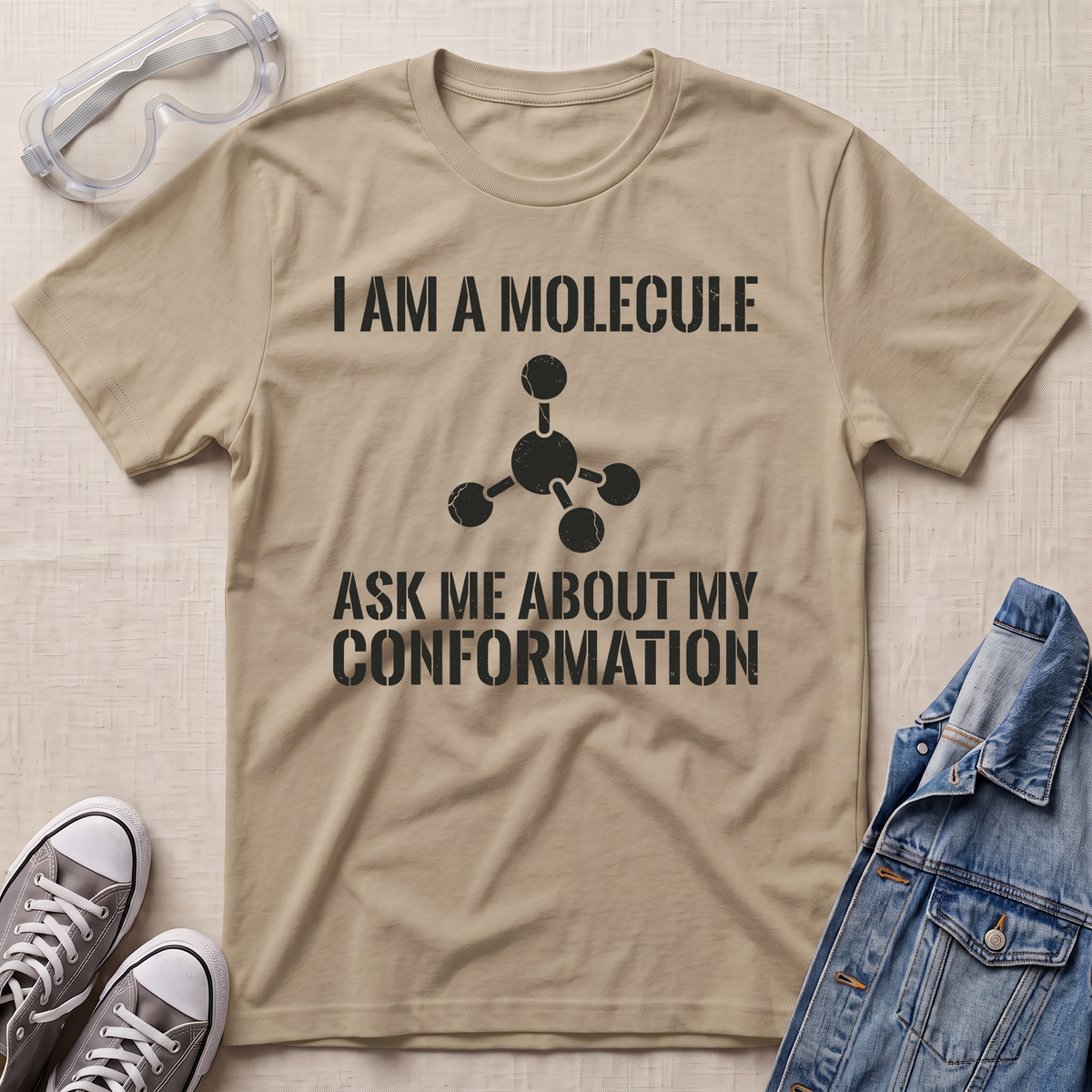 A beige graphic t-shirt laid flat on a textured beige background, styled with safety goggles, gray sneakers, and a blue denim jacket. The t-shirt features a black printed graphic of a molecule and the text 'I AM A MOLECULE ASK ME ABOUT MY CONFORMATION'.