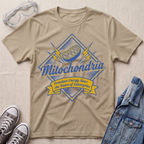 A beige graphic t-shirt featuring a blue and yellow 'Mitochondria' design, styled in a flat lay with safety goggles, gray sneakers, and a denim jacket on a textured beige fabric background.
