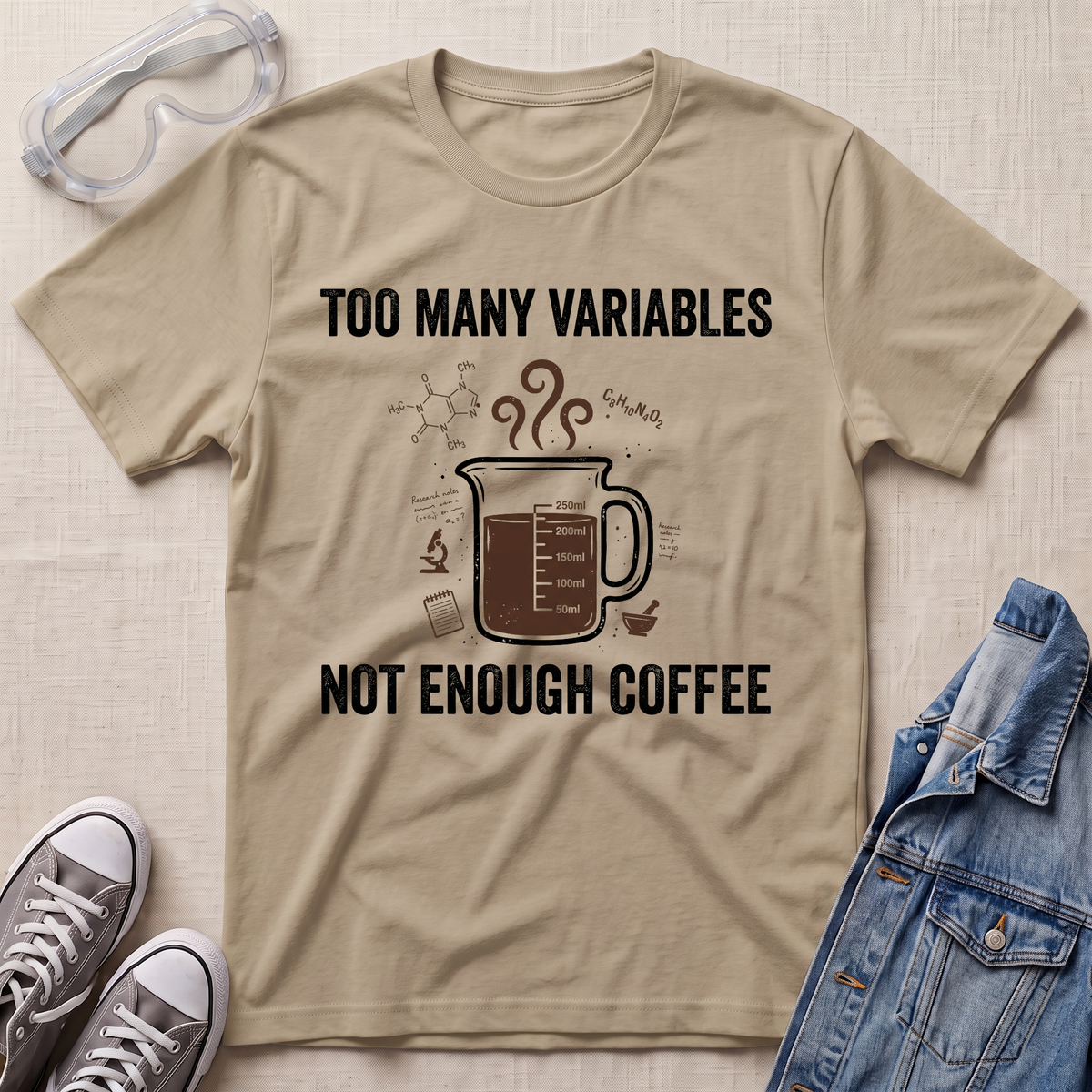A beige graphic t-shirt laid flat on a textured fabric background, styled with safety goggles, gray sneakers, and a blue denim jacket. The t-shirt features a dark brown coffee mug illustration with the text 'TOO MANY VARIABLES NOT ENOUGH COFFEE'.