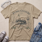 A beige graphic t-shirt featuring a vintage-style illustration of an ant with the text 'Fungi Culture' and 'Spreading Puddles and Spores'. The t-shirt is styled in a flat-lay arrangement with safety goggles, gray sneakers, and a blue denim jacket on a textured beige fabric background.