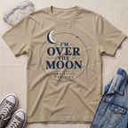 A sand graphic t-shirt laid flat on a textured light beige background, surrounded by styled props including clear safety goggles, gray sneakers, and a blue denim jacket. The t-shirt features a navy blue graphic of a crescent moon inside a circle with the text 'I'M OVER THE MOON AND STILL ORBITING'.