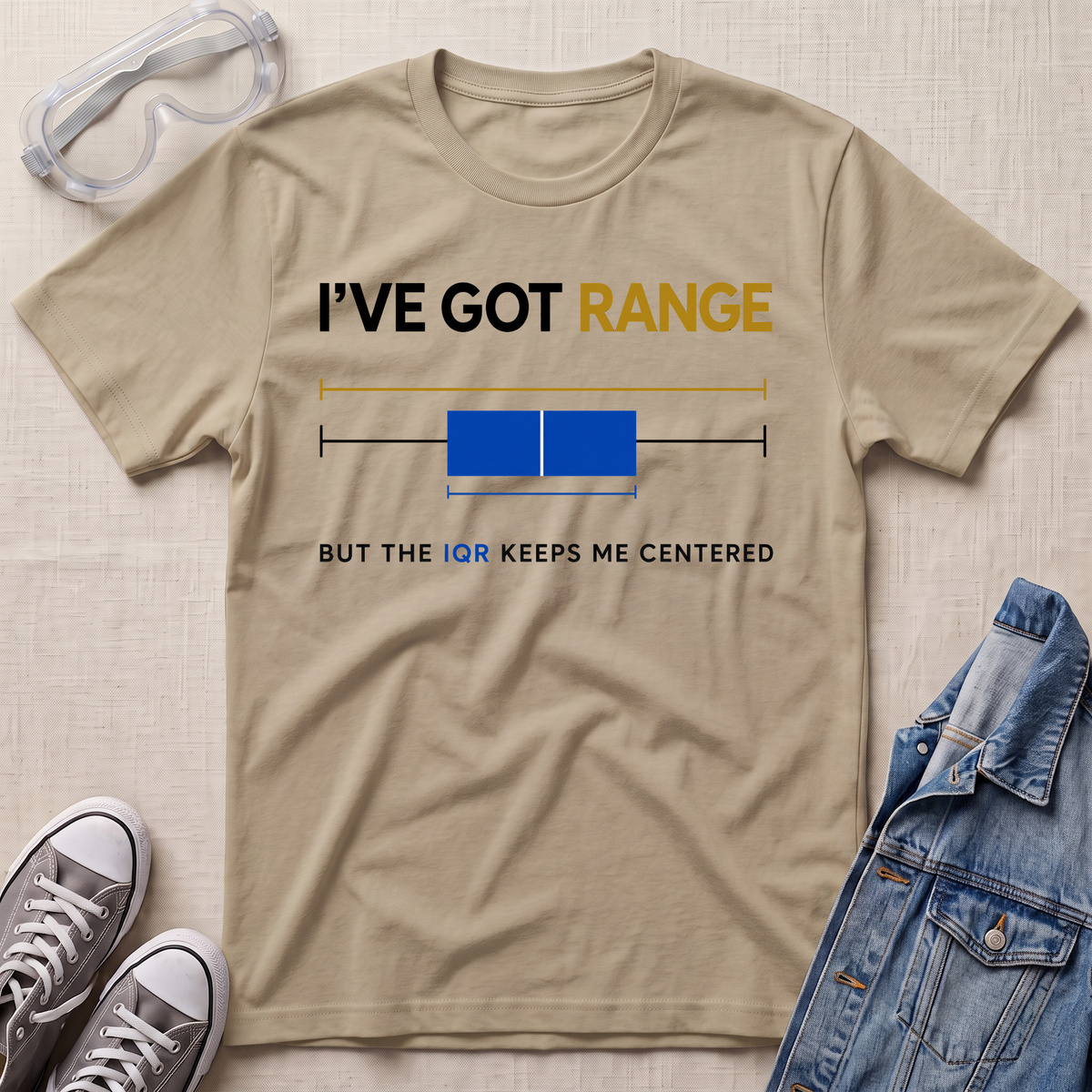 A beige graphic t-shirt laid flat on a textured light beige surface, styled with safety goggles, gray sneakers, and a blue denim jacket. The t-shirt features a humorous sci-fi graphic with the text 'I'VE GOT RANGE BUT THE IQR KEEPS ME CENTERED' and a simple blue and yellow box plot illustration.