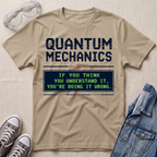 A beige graphic T-shirt laid flat on a light fabric surface, styled with safety goggles, gray sneakers, and a denim jacket. The shirt has a large dark blue 'QUANTUM MECHANICS' text and a green text box reading, 'IF YOU THINK YOU UNDERSTAND IT, YOU'RE DOING IT WRONG.