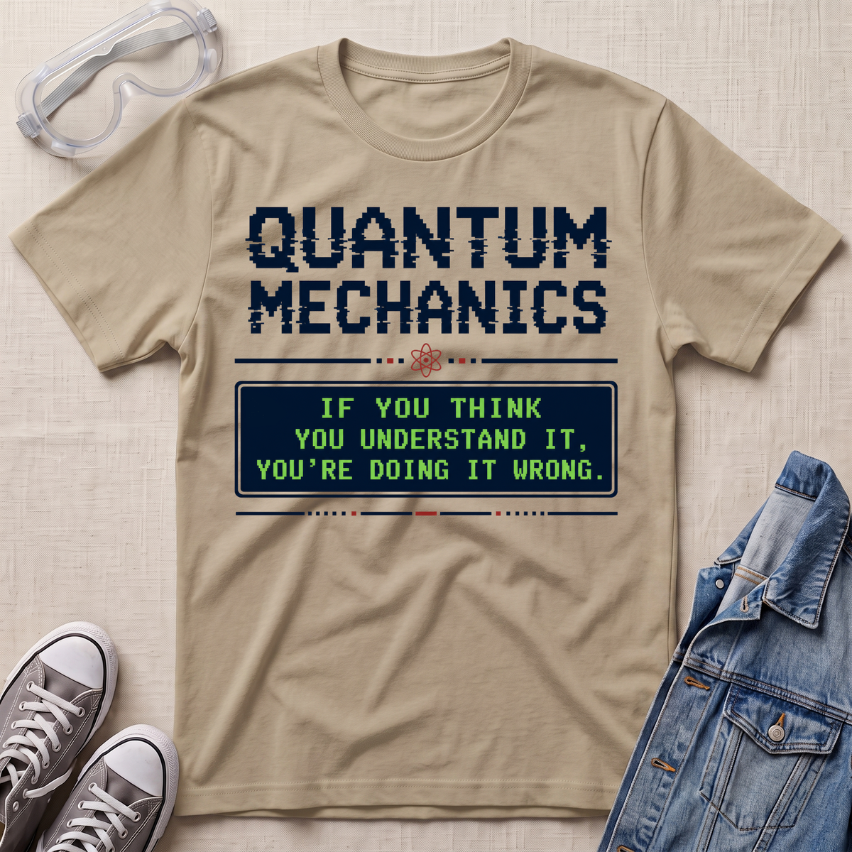 A beige graphic T-shirt laid flat on a light fabric surface, styled with safety goggles, gray sneakers, and a denim jacket. The shirt has a large dark blue 'QUANTUM MECHANICS' text and a green text box reading, 'IF YOU THINK YOU UNDERSTAND IT, YOU'RE DOING IT WRONG.