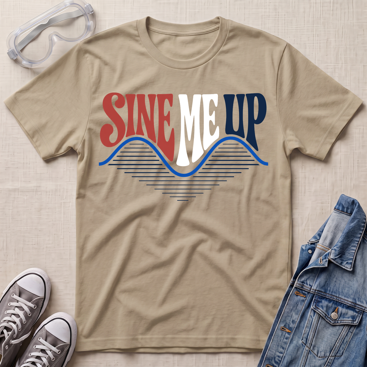 A beige graphic t-shirt laid flat on a textured light beige surface, styled with clear safety goggles, gray sneakers, and a blue denim jacket. The t-shirt features a retro graphic reading 'SINE ME UP' in red, white, and blue, with a wavy blue line design underneath.