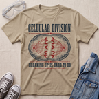 A beige graphic t-shirt laid flat on a textured fabric background, styled with safety goggles, gray sneakers, and a blue denim jacket. The t-shirt features a distressed graphic of a dividing cell with the text 'CELLULAR DIVISION' and 'BREAKING UP IS HARD TO DO'.