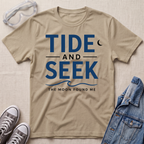 A sand beige short-sleeve graphic T-shirt laid flat on a light beige textured surface, with safety glasses, gray sneakers, and a folded denim jacket around it. The shirt has large navy text reading 'TIDE AND SEEK' with smaller text 'THE MOON FOUND ME' and a crescent moon graphic.