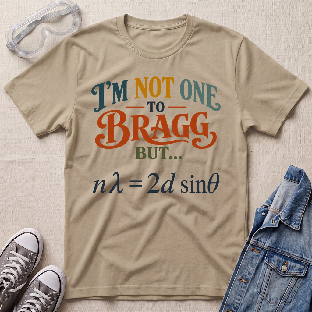 A beige graphic t-shirt laid flat on a textured fabric background, styled with safety goggles, gray sneakers, and a blue denim jacket. The t-shirt features the text 'I'M NOT ONE TO BRAGG BUT.