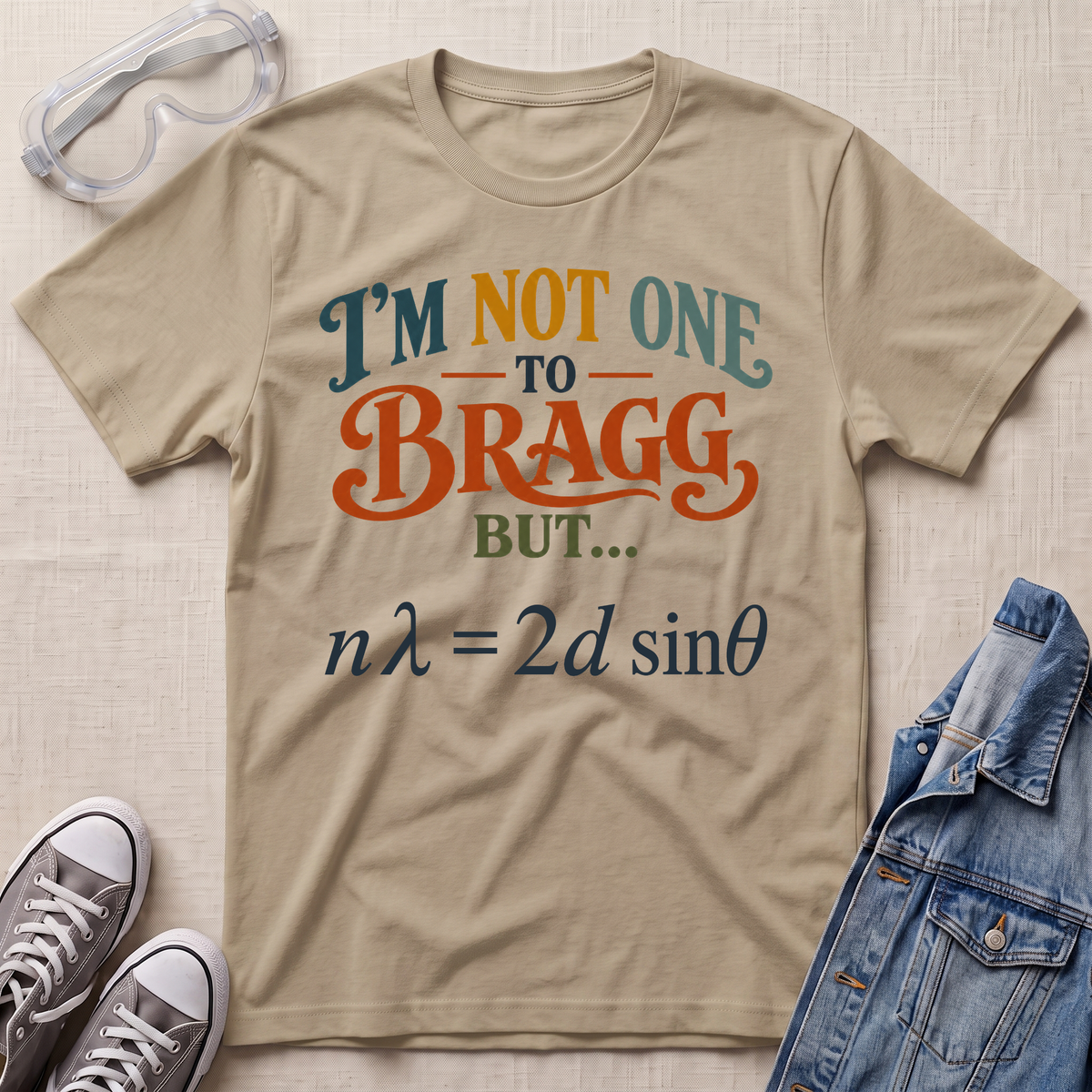 A beige graphic t-shirt laid flat on a textured fabric background, styled with safety goggles, gray sneakers, and a blue denim jacket. The t-shirt features the text 'I'M NOT ONE TO BRAGG BUT.