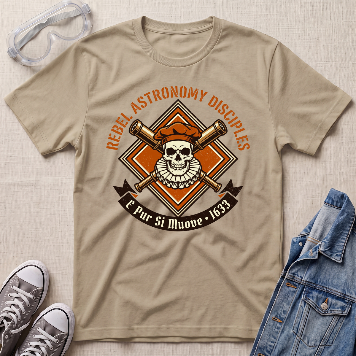 A beige graphic t-shirt laid flat on a textured fabric background, styled with safety goggles, gray sneakers, and a blue denim jacket. The t-shirt features a retro-style astronomy-themed graphic with a skull, crossed telescopes, and the text 'REBEL ASTRONOMY DISCIPLES' and 'E Pur Si Muove - 1633'.
