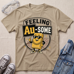 A beige graphic t-shirt laid flat on a textured fabric background, styled with safety goggles, gray sneakers, and a denim jacket. The t-shirt features a cartoon gold bar character wearing sunglasses and a gold chain, with the text 'FEELING Au-SOME' in a circular badge design.