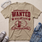 A beige graphic t-shirt featuring Schrödinger's Cat in a 'WANTED' poster design, styled in a flat-lay with a denim jacket, sneakers, and safety goggles on a textured beige background.