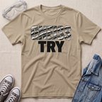 A beige graphic t-shirt laid flat on a textured light beige background, styled with safety goggles, gray sneakers, and a blue denim jacket. The t-shirt features the text 'GNEISS TRY' with 'GNEISS' filled with a black and white camouflage pattern and 'TRY' in solid black below it.
