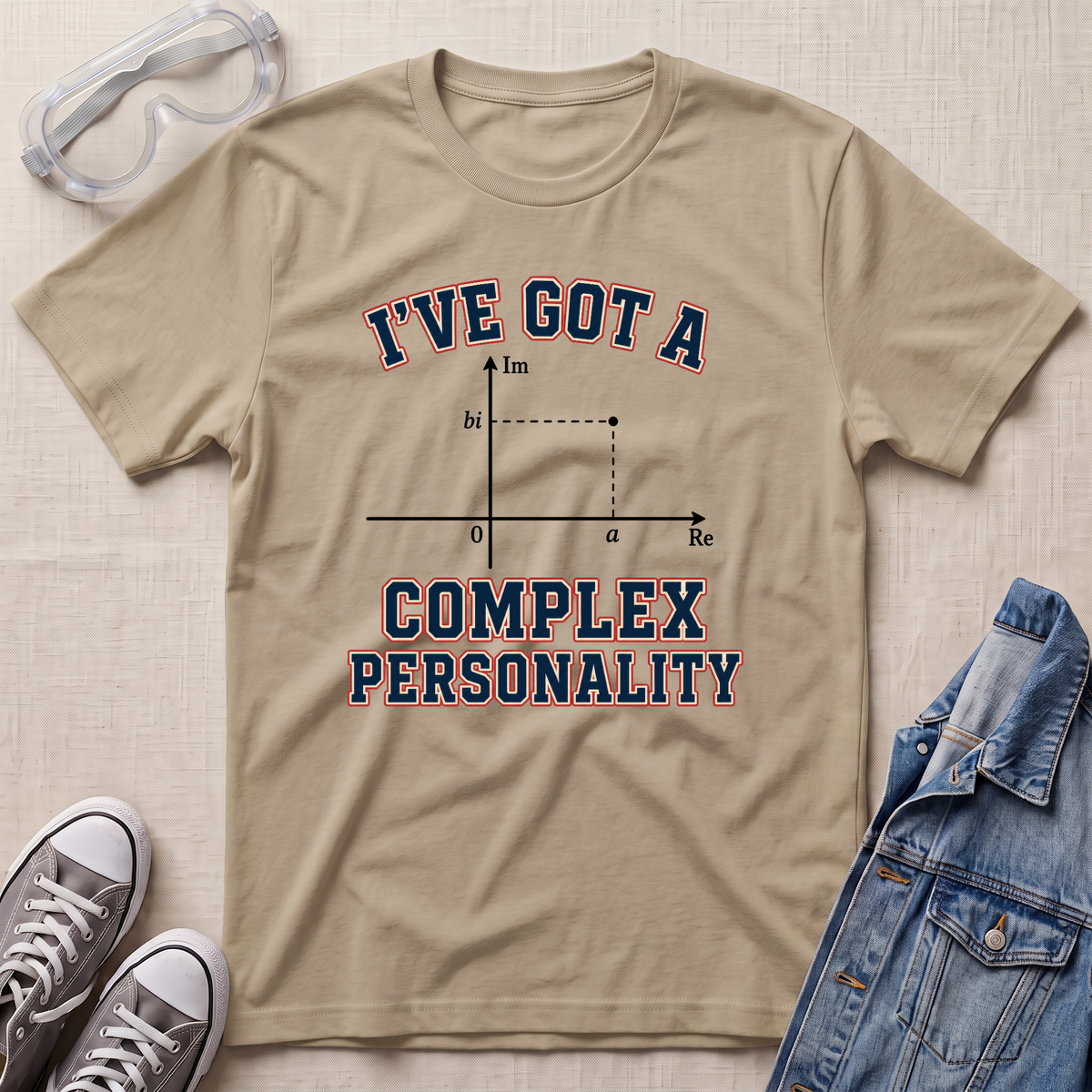 A beige graphic t-shirt laid flat on a textured beige background, styled with safety goggles, gray sneakers, and a blue denim jacket. The t-shirt features a complex number plot graphic with the text 'I'VE GOT A COMPLEX PERSONALITY' in navy blue with a red outline.