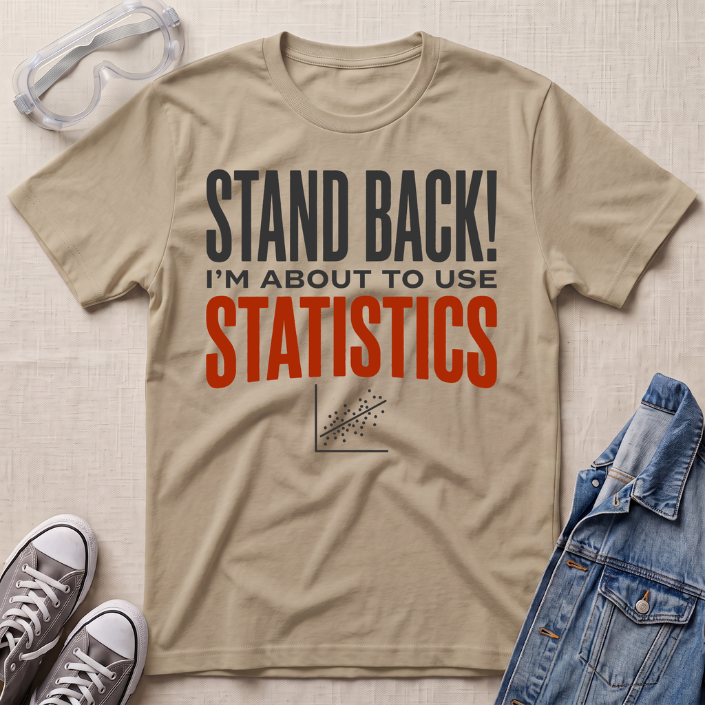 A beige graphic t-shirt laid flat on a textured beige background, surrounded by props including safety goggles, gray sneakers, and a blue denim jacket. The t-shirt features the text 'STAND BACK! I'M ABOUT TO USE STATISTICS' in dark gray and orange-red lettering.
