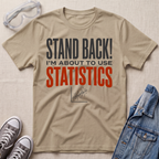 A beige graphic t-shirt laid flat on a textured beige background, surrounded by props including safety goggles, gray sneakers, and a blue denim jacket. The t-shirt features the text 'STAND BACK! I'M ABOUT TO USE STATISTICS' in dark gray and orange-red lettering.