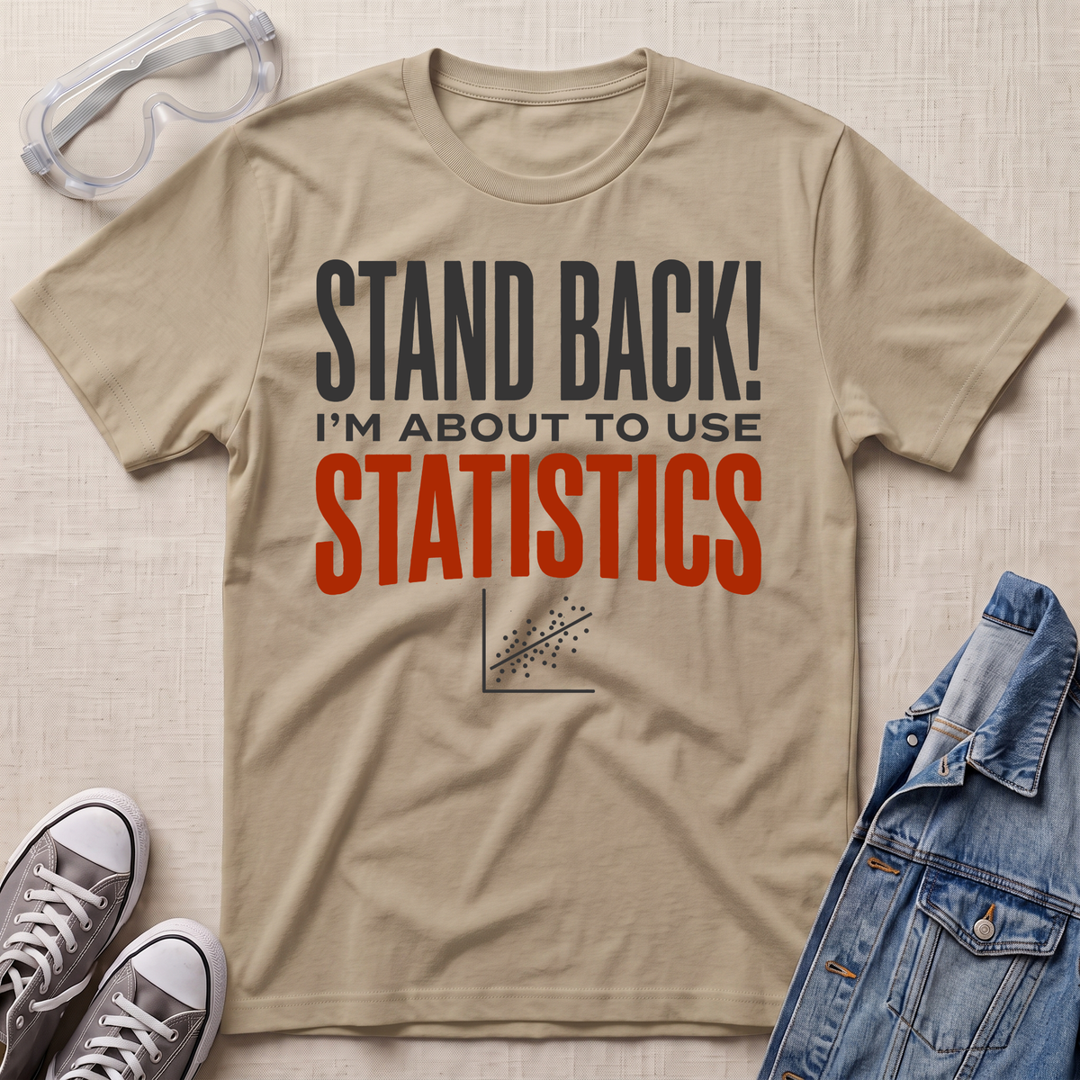 A beige graphic t-shirt laid flat on a textured beige background, surrounded by props including safety goggles, gray sneakers, and a blue denim jacket. The t-shirt features the text 'STAND BACK! I'M ABOUT TO USE STATISTICS' in dark gray and orange-red lettering.