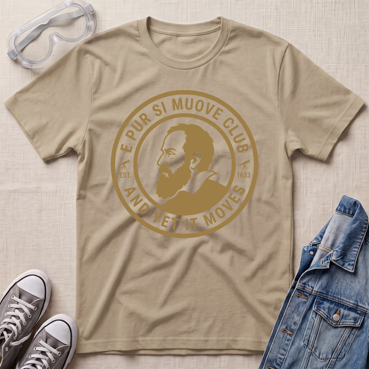 A sand beige short-sleeve graphic T-shirt laid flat on a light beige textured surface, surrounded by styling props including clear safety goggles, gray sneakers, and a blue denim jacket. The shirt features a large circular tan/gold print with a portrait and the text 'E PUR SI MUOVE CLUB AND YET IT MOVES'.
