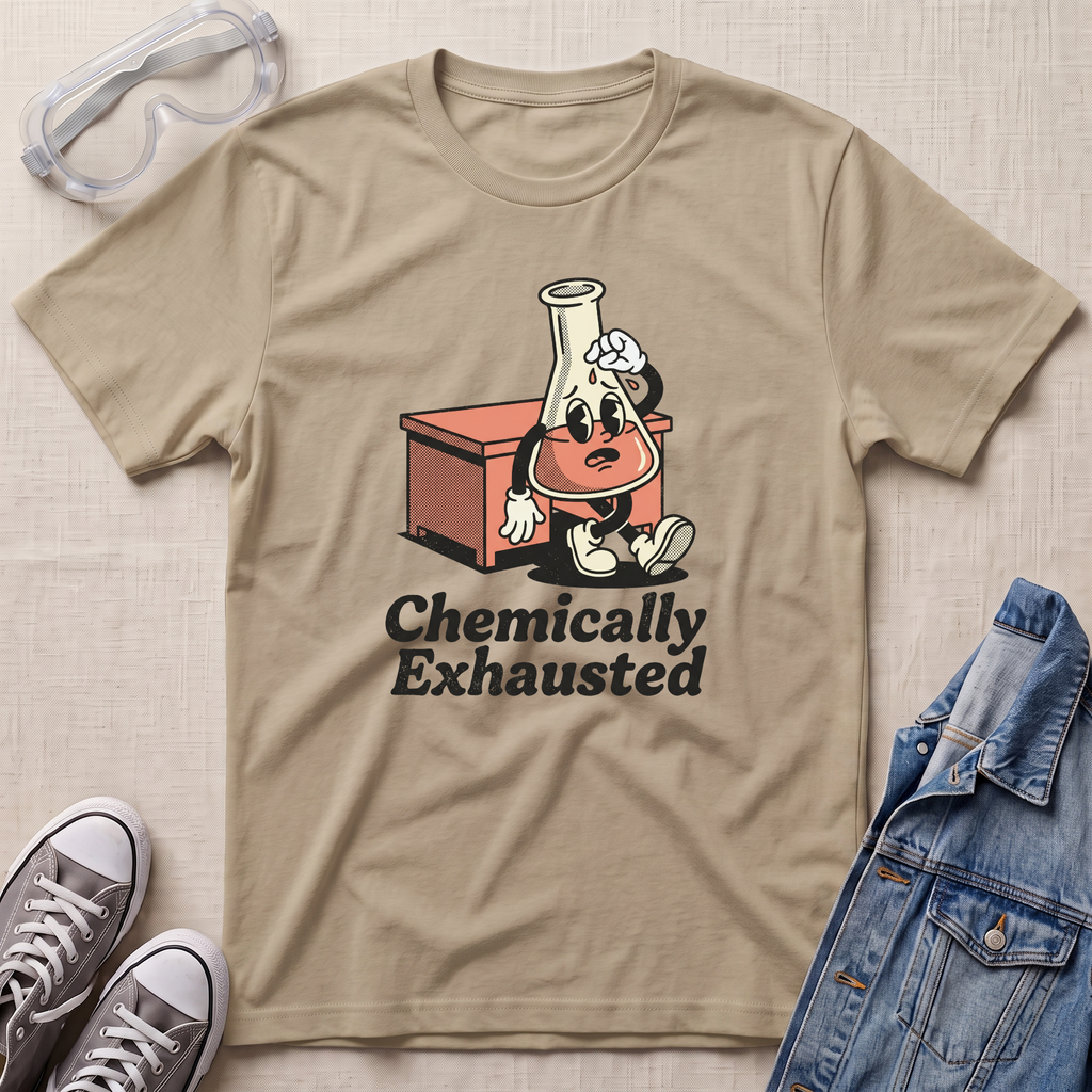 A beige graphic t-shirt laid flat on a textured fabric background, styled with safety goggles, gray sneakers, and a blue denim jacket. The t-shirt features a cartoon graphic of a chemist character sitting next to an orange beaker and the text 'Chemically Exhausted'.