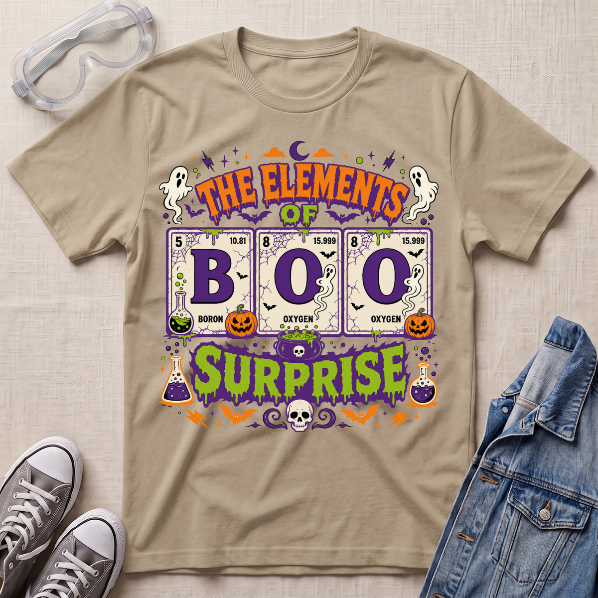 A beige graphic T-shirt laid flat on a light fabric background, styled with clear safety goggles, gray sneakers, and a denim jacket. The shirt features a colorful Halloween-themed design reading 'THE ELEMENTS OF BOO SURPRISE' with periodic-table-style elements, ghosts, pumpkins, bats, and purple decorative accents.