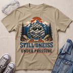 A beige graphic t-shirt laid flat on a textured fabric background, styled with safety goggles, gray sneakers, and a blue denim jacket. The t-shirt features a graphic of a gneiss rock wearing sunglasses with mountain, trees, and an orange sun in the background, with the text 'STILL GNEISS UNDER PRESSURE'.