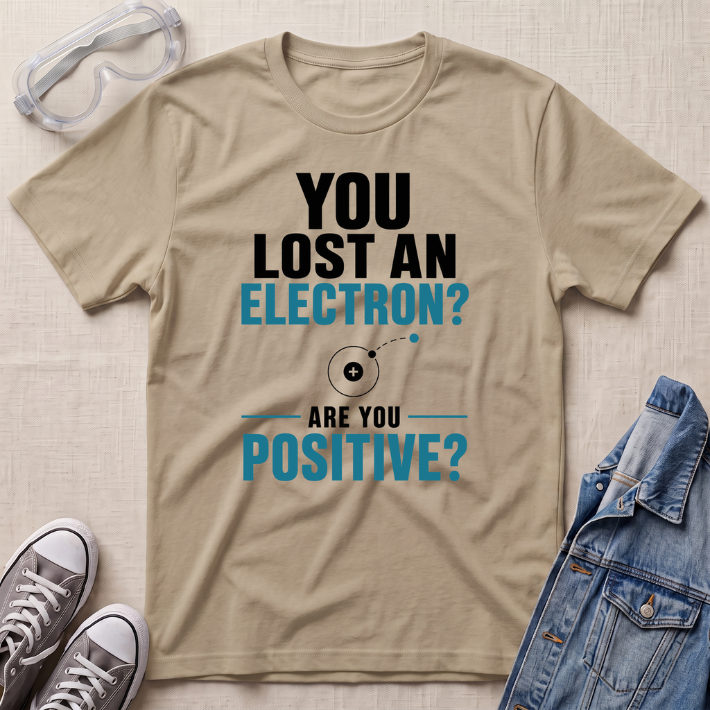 A beige graphic t-shirt laid flat on a textured beige background, surrounded by styling props including safety goggles, gray sneakers, and a blue denim jacket. The t-shirt features a black and teal text graphic that reads 'YOU LOST AN ELECTRON? ARE YOU POSITIVE?' with a simple atom illustration.