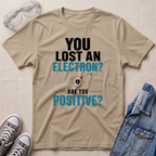 A beige graphic t-shirt laid flat on a textured beige background, surrounded by styling props including safety goggles, gray sneakers, and a blue denim jacket. The t-shirt features a black and teal text graphic that reads 'YOU LOST AN ELECTRON? ARE YOU POSITIVE?' with a simple atom illustration.