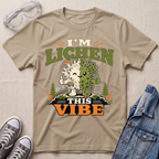 A beige science t-shirt featuring a cute lichen character with the pun “I’m Lichen This Vibe,” surrounded by retro botanical and nature-inspired accents.