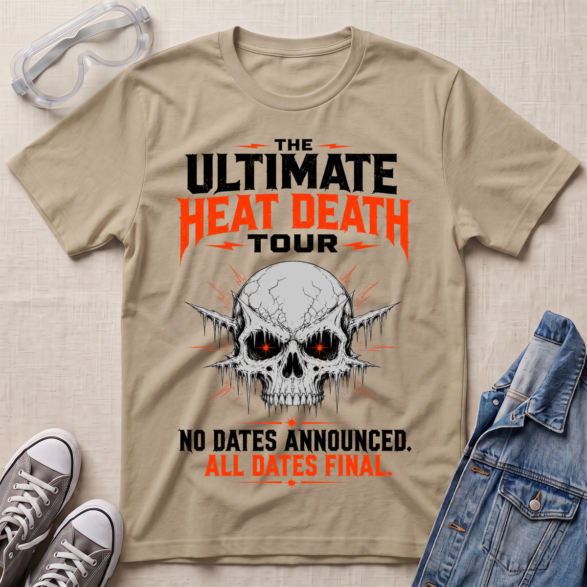 A beige graphic t-shirt laid flat on a textured fabric background, styled with safety goggles, gray sneakers, and a denim jacket. The t-shirt features a skull graphic with glowing red eyes and text that reads 'THE ULTIMATE HEAT DEATH TOUR' and 'NO DATES ANNOUNCED.