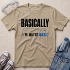 A beige graphic t-shirt laid flat on a textured light background, styled with safety goggles, gray sneakers, and a blue denim jacket. The t-shirt features the text 'BASICALLY I'M QUITE BASIC' in black and blue lettering.