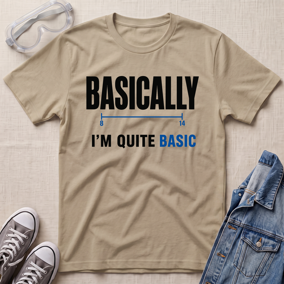 A beige graphic t-shirt laid flat on a textured light background, styled with safety goggles, gray sneakers, and a blue denim jacket. The t-shirt features the text 'BASICALLY I'M QUITE BASIC' in black and blue lettering.