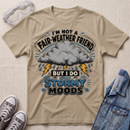 A beige graphic t-shirt laid flat on a textured fabric background, styled with safety goggles, gray sneakers, and a blue denim jacket. The t-shirt features a storm graphic with a gray cloud, lightning bolts, and the text 'I'M NOT A FAIR-WEATHER FRIEND BUT I DO HAVE STORMY MOODS'.